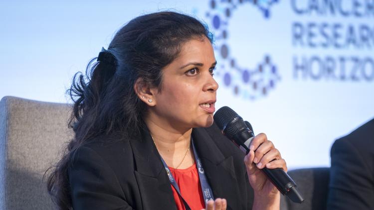 A woman sat on a stage speaking into a microphone, with the Cancer Research Horizons logo behind her