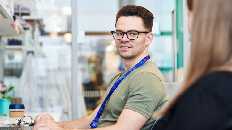 A person sat at a desk, short hair, glasses and short sleeved shirt wearing a lanyard, looking at the person next to them