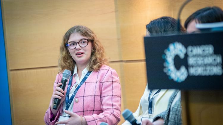 A young woman holding a microphone. She is wearing a pink blazer and glasses. She has blonde medium-length wavy hair.  