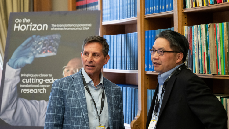 Two men stand in a library setting. The man on the left wears a blue checked blazer, while the man on the right wears glasses and a dark jacket. Both have lanyards. Bookshelves are visible in the background.