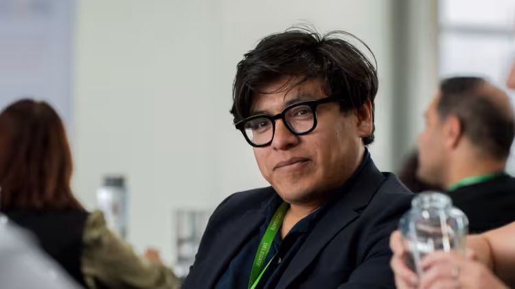 A person with dark short hair and a black shirt, wearing glasses and a lanyard in conversation at a networking event.