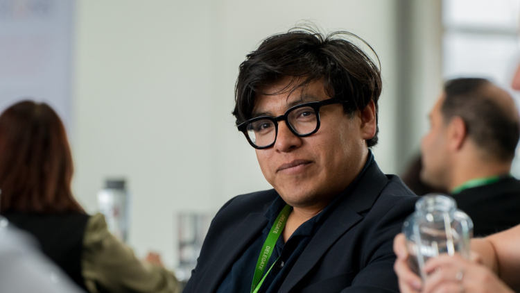 A person with dark short hair and a black shirt, wearing glasses and a lanyard in conversation at a networking event.