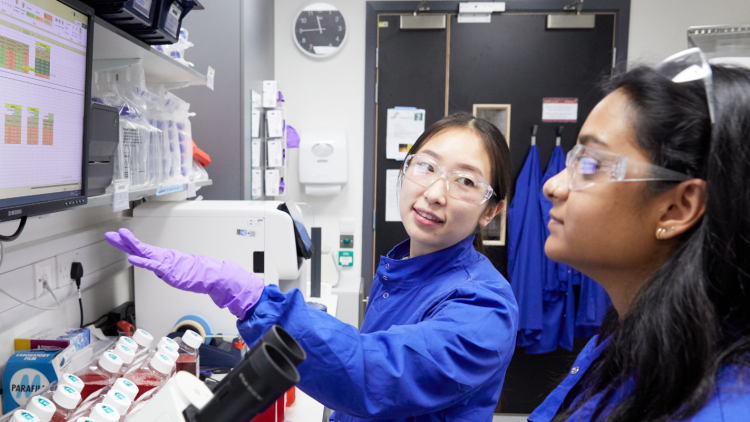 Two scientists in blue lab coats pointing at equipment.