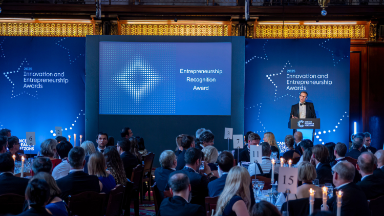 A person giving a speech to the Awards ceremony audience, in front of a blue backdrop with the event name on it. 