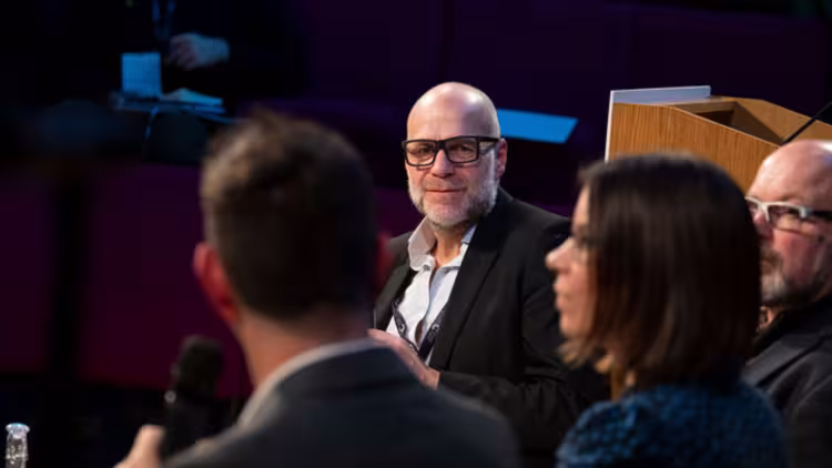 A person participating in a panel at the Innovation Summit. They wear a white shirt, black blazer and dark rimmed glassed