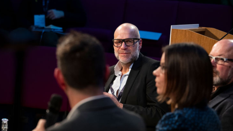 A person participating in a panel at the Innovation Summit. They wear a white shirt, black blazer and dark rimmed glassed