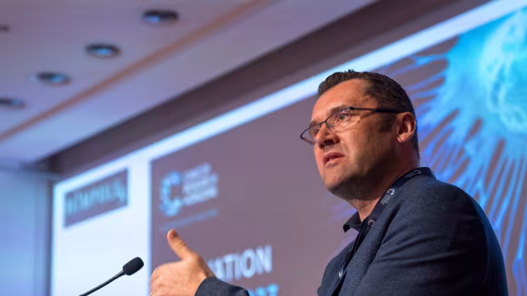 A person wearing glasses, short dark hair and a dark blazer giving a talk at the Innovation Summit
