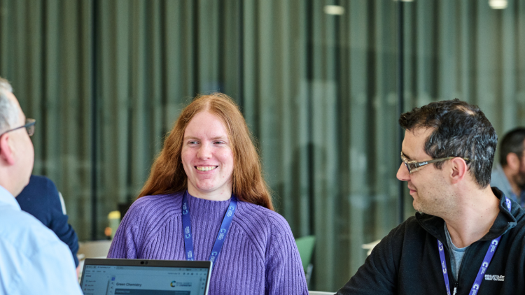 Two researchers having a meeting. One of them has long, red hair and wears a purple jumper. The other has dark, short hair and wears a black t-shirt. 