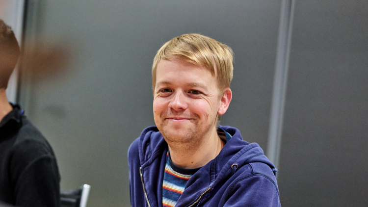 A person with short, blonde hair and a dark blue hoodie smiling in a meeting. 